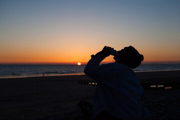 Silhouette of a woman drinking from a can in front of the setting sun.