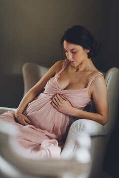 Pregnant woman sits on armchair wearing a beautiful pale pink dress