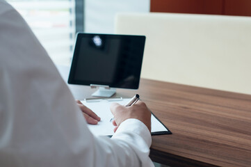 Businessman in a white shirt with a pen in his hands signs a contract in the office. Workplace with document and digital tablet on a wooden table.