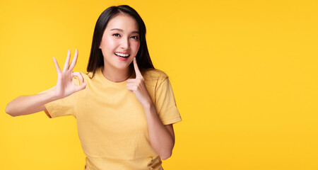 Cheerful young woman using finger point to her whitening teeth while make hand sign ok over isolated yellow background. She showing her cleaning fresh toothy. Dental care and healthy concept.