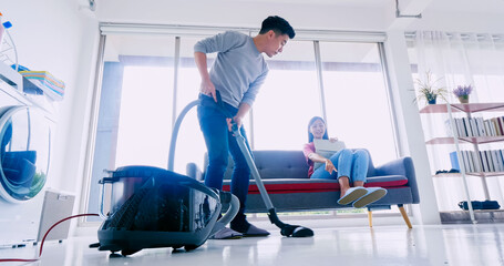 Husband cleaning floor with vacuum cleaner while wife working with laptop on sofa. Happy Asian couple spending time together at home.