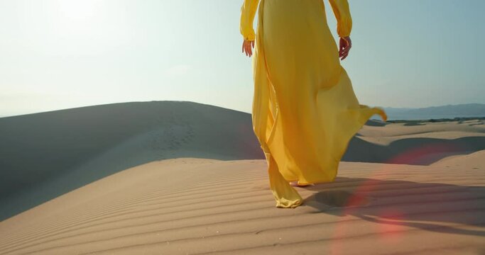 Beautiful fashion video. Bright dress is flying by the wind in slow motion with blue sky on background. Commercial footage for clothing business. Cinematic modern shot of woman walking by desert sand
