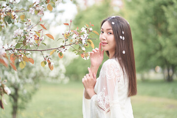 Portrait of a beautiful Asian woman in a white dress, standing near a tree with white flowers, holding her hands near her face and looking directly at the camera