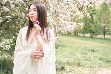 Portrait of a beautiful Asian woman in a white dress, she is standing near a tree with white flowers, on her cheeks flowers with petals are glued on a band-aid, she looks away