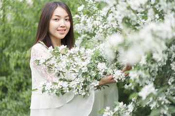Portrait of a beautiful Asian woman in a white dress, standing near a tree with white flowers, holding branches with her hands and looking directly at the camera