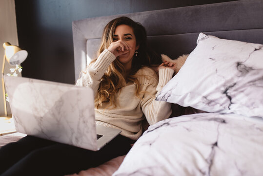 A young woman using her laptop in bed with her dog next to her
