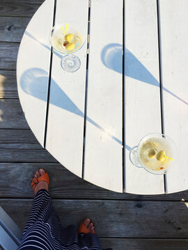 Two martinis casting long shadows sit on a round, grey table in summer.