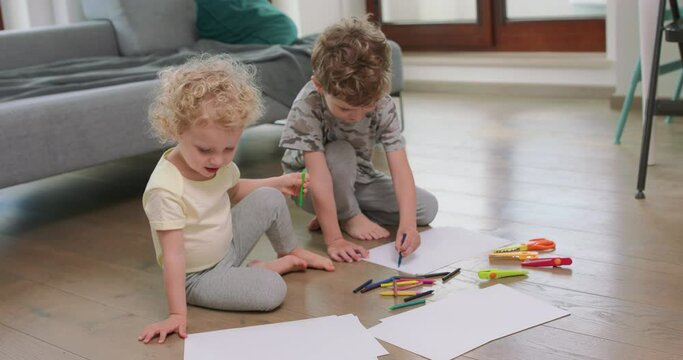 A little boy and a little girl are drawing with pencils on on the white paper on the floor. Looking at each other from time to time. Pencils, paper, kid's scissors and paper knives are in front of