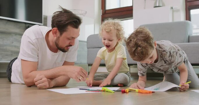A little boy and a little girl, and their father are drawing with pencils on the white paper on the floor. Father talks to girl. Pencils, paper, kid's scissors and paper knives are in front of them