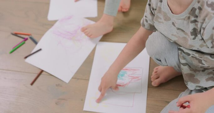 Close up of a hands and feet of a little boy and a little girl, who are drawing with pencils on the white paper on the floor. Pencils and paper are in front of them.