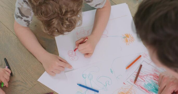 Close up of a little boy's and a little girl's, and their father's heads and hands. They are drawing with pencils on the white paper on the floor. Boy's picture focused.