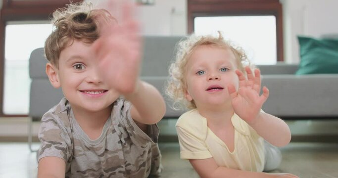 Close up of a little boy and a little girl, who are laying on the floor and waving hands, looking to the camera. Grey sofa and windows in the background.