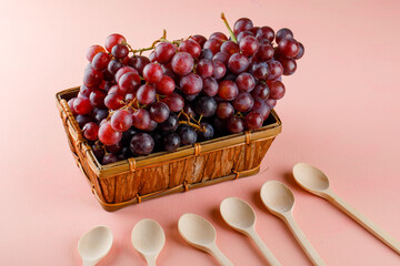 Red grapes with wooden spoons in a basket on pink background, high angle view.