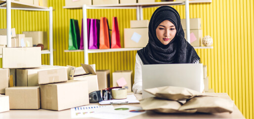 Portrait of smiling beautiful muslim owner asian woman freelancer sme business online shopping working on laptop computer with parcel box on table at home - Business online shipping and delivery