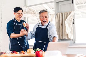 Portrait of happy love asian family senior mature father and young adult son having fun cooking together and looking for recipe on Internet with laptop computer to prepare the yummy eating lunch 
