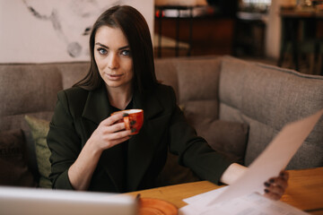 Serious young business woman is working with paper documents in cafe sitting at table with laptop ,looking at the camera. Lady enjoys delicious coffee while working. 