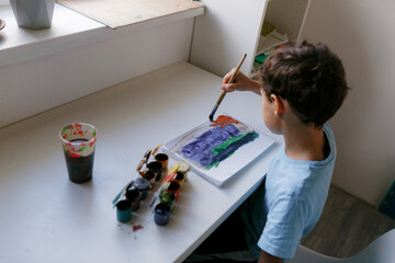 Fototapeta na wymiar Curly dark-haired boy in a blue T-shirt draws a picture with paints while sitting at a white table
