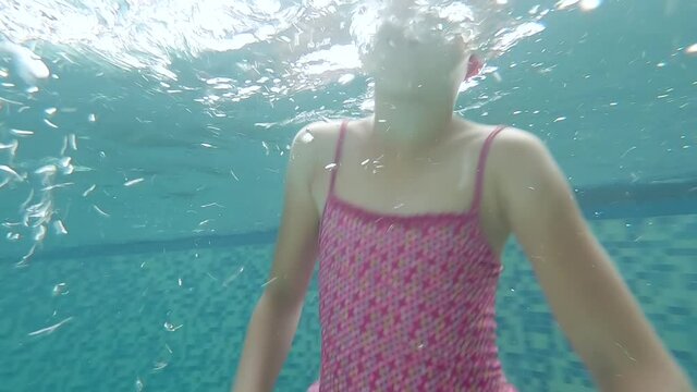 Funny little girl fooling around in the pool underwater and smiling. A child in a red bathing suit, cap and goggles dives and swims