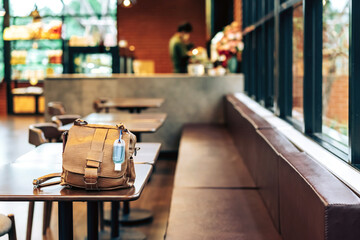 Mini portable alcohol gel bottle to kill Corona Virus(Covid-19) hang on a brown leather shoulder bag on table in coffee shop.New normal lifestyle. Health care concept. Selective focus on alcohol gel