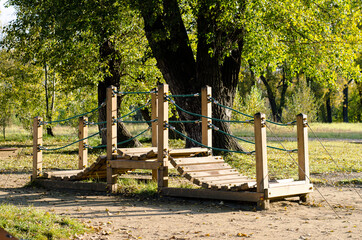 Wooden play bridge for children, located in the Park among the trees. Outdoor play facilities. Bridge made of wooden planks with rope railings.