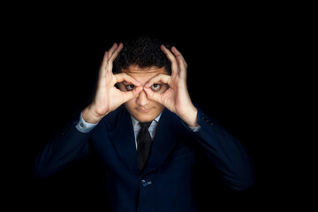 Frustrated man making a pair of glasses and giving a humours owl expression isolated on black background. Man in suit giving humor gesture.