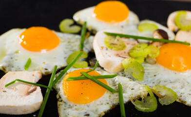 Closeup of fried quail eggs with sliced raw champignons, celery and green onion on black background