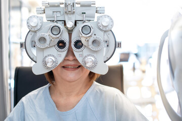 Senior Asian woman looking through optical phoropter during eye exam, diagnostic ophthalmology equipment, selective focus