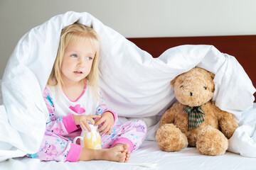 Obraz na płótnie Canvas Cute little girl with blonde hair sitting on the bed with stuffed teddy bear. Happy childhood. Stay at home during coronavirus covid-19 pandemic quarantine concept.