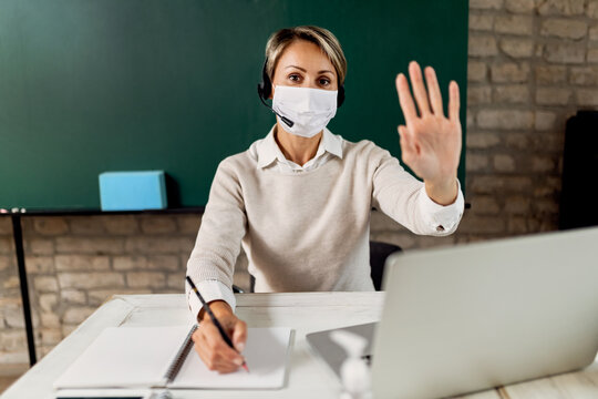 Female Teacher With Face Mask Taking Notes And Waving From The Classroom.