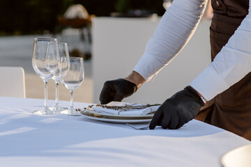 Obraz na płótnie Canvas waiters hands in protective black gloves arrange a wedding party reception table decorated with flowers: plates, forks, knives and wine glasses.