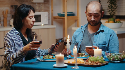 Focused married couple reading news of phones during romantic dinner. Adults sitting at the table, browsing online, surfing, using smartphones, celebrating their anniversary in the dining room.