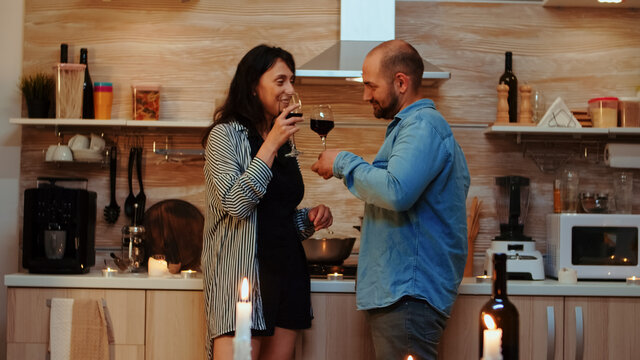 Young caucasian couple having romantic date at home in the kitchen, drinking red wine, talking, smiling in dining room. Two people in love having pleasant conversation during healthy meal.