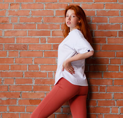 Fashion style studio portrait of beautiful red-haired young girl in leggings pants and white t-shirt. Model standing and posing beside brick wall