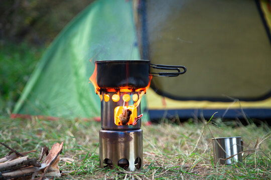 food is cooked on a camping wood stove. green tent on the grass at the background, firewoods and steel mug near the wood stove