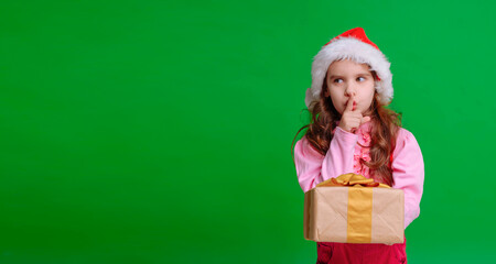 Cute girl in a Santa hat holds a Christmas gift isolated on a green background.