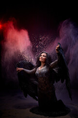 Girl in black dress and with black wings posing in dark studo during photoshoot with flour or dust and light. A fallen angel begs and asks for something
