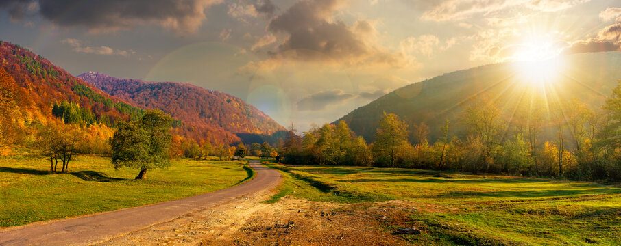 road winding through the country valley at sunset. wonderful autumn landscape in mountains in evening light. forest on hills in colorful foliage. sunny weather with fluffy clouds on the sky