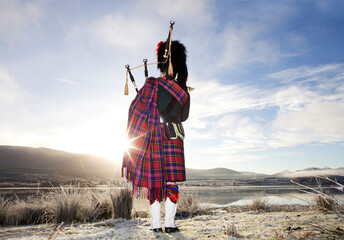 Scottish Bagpiper playing bagpipes by loch at sunrise. Scotland.