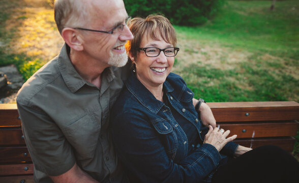 Happy middle aged, retired couple laughing and cuddling outside sitting on a bench