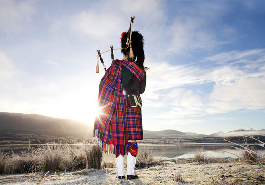 Scottish Bagpiper playing bagpipes by loch at sunrise. Scotland.