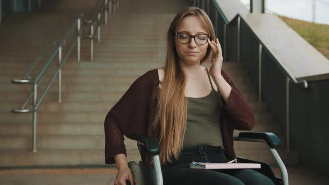 Young discouraged caucasian woman with sad face expression, in the wheelchair in front of the staircase looking around for accessible way. High quality 4k footage