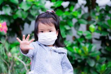 Cute Asian girl wearing a white medical face mask. Raise your hand as a sign to stop. The child looks at the camera. Children take social distancing seriously. Little kid in gray shirt, 3 years old.