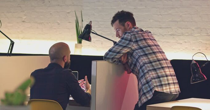 Wide shot of two co-workers talking over their workspace to each other.