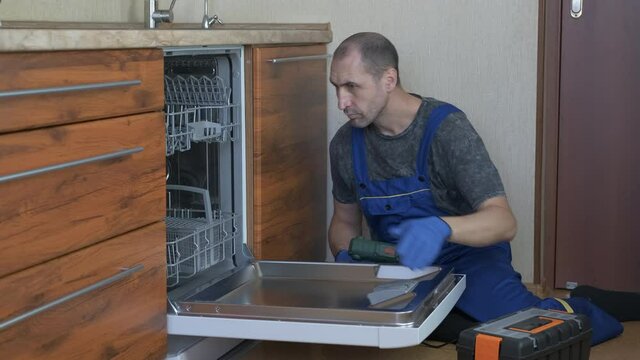 Repair technic in the kitchen. A happy man with a drill open the door jf the dishwashing machine and repair it.