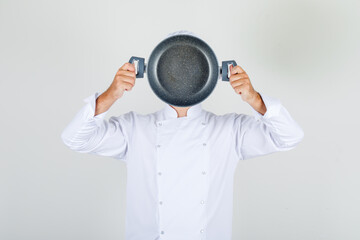 Male chef holding empty pan over his face in white uniform front view.