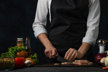 The chef in white shirt and black apron cuts meat on the professional kitchen. Vegetables, mushrooms and spices are background. The girl's hands with knife. Food concept.