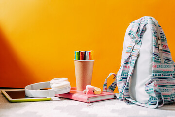 School supplies on the orange background. Blue backpack, white headphones, notebook and pens, tablet.