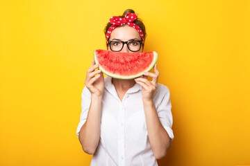 Young woman in a red headband and glasses covers half of her face with watermelon on a yellow background