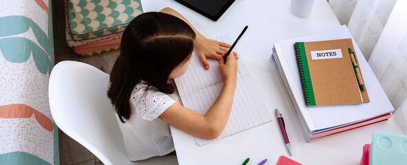 Top view of little girl studying at home with tablet and mask on table
