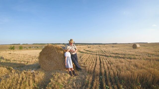 Little girl with grandfather in field haystacks, grandfather farmer is teaching the younger generation using a tablet. The concept of generations, technology and farming.